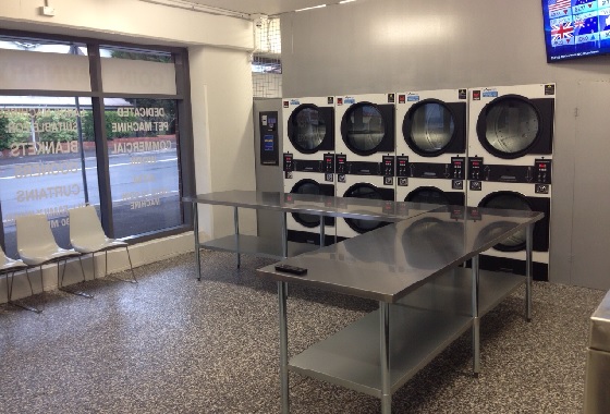 Inside Katoomba Laundromat: a row of stacked commercial washers and dryers beside stainless steel folding benches, with seating by the front window.